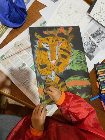 A young artist wearing a red smock works on a colorful tiger artwork using oil pastels on black paper, surrounded by sketch references and photos of real tigers on a wooden table.