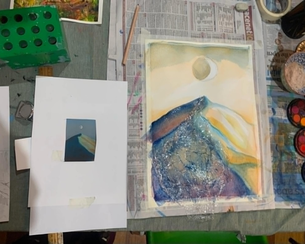 A work-in-progress mountain landscape painting displayed on an art table beside a printed reference image. Watercolor and mixed-media techniques create layered blue, purple, and golden tones, with art supplies, newspapers, and paint palettes surrounding the artwork in a studio setting.