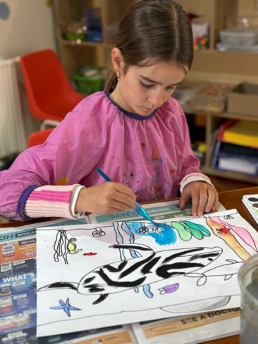 Young girl wearing a pink art smock draws and colours a large shark illustration with bright blue, green, pink, and purple details at a table in an art studio.